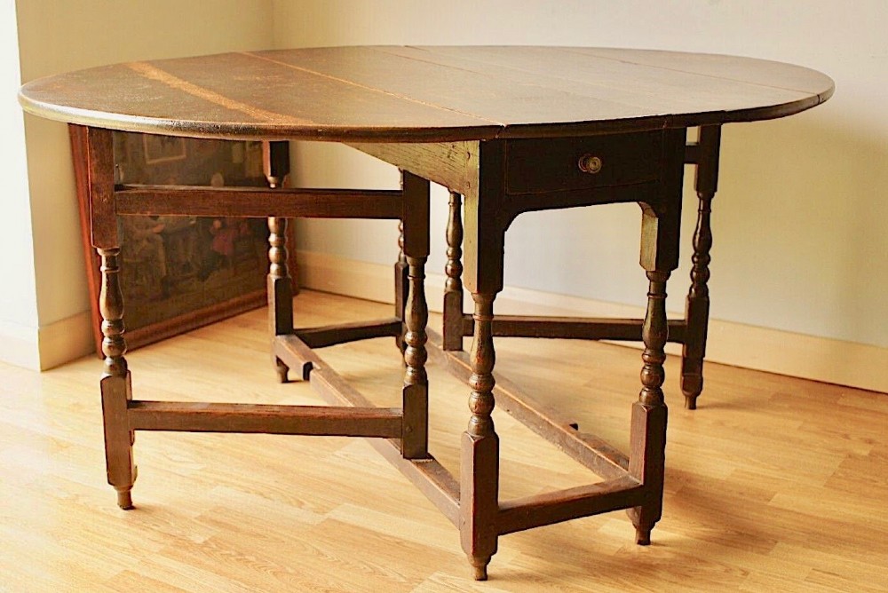 A dark oak Georgian gateleg dining table featuring two oval drop leaves, turned legs, a lower stretcher, and a single end drawer.