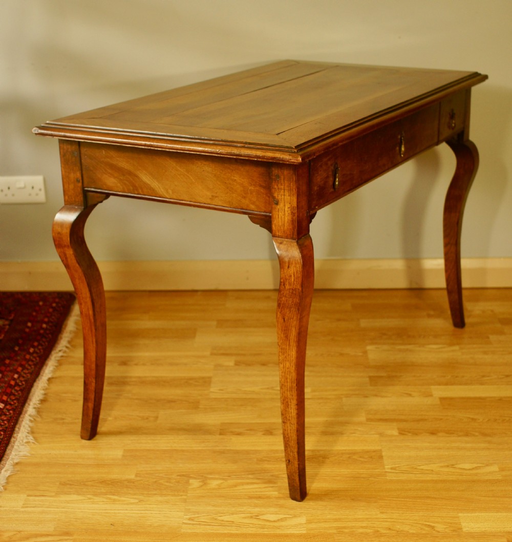 An antique French walnut writing table featuring three frieze drawers, brass handles, and cabriole legs against a carved wooden panelled backdrop. View IV