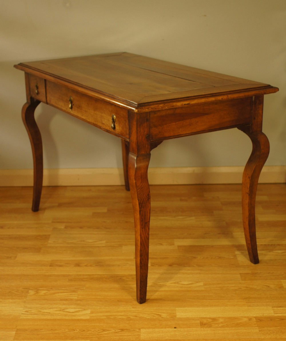 An antique French walnut writing table featuring three frieze drawers, brass handles, and cabriole legs against a carved wooden panelled backdrop. View V