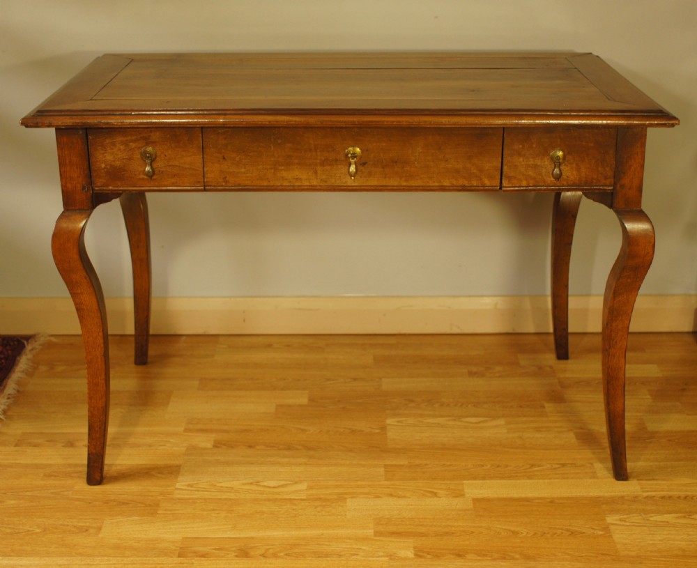 An antique French walnut writing table featuring three frieze drawers, brass handles, and cabriole legs against a carved wooden panelled backdrop. View II