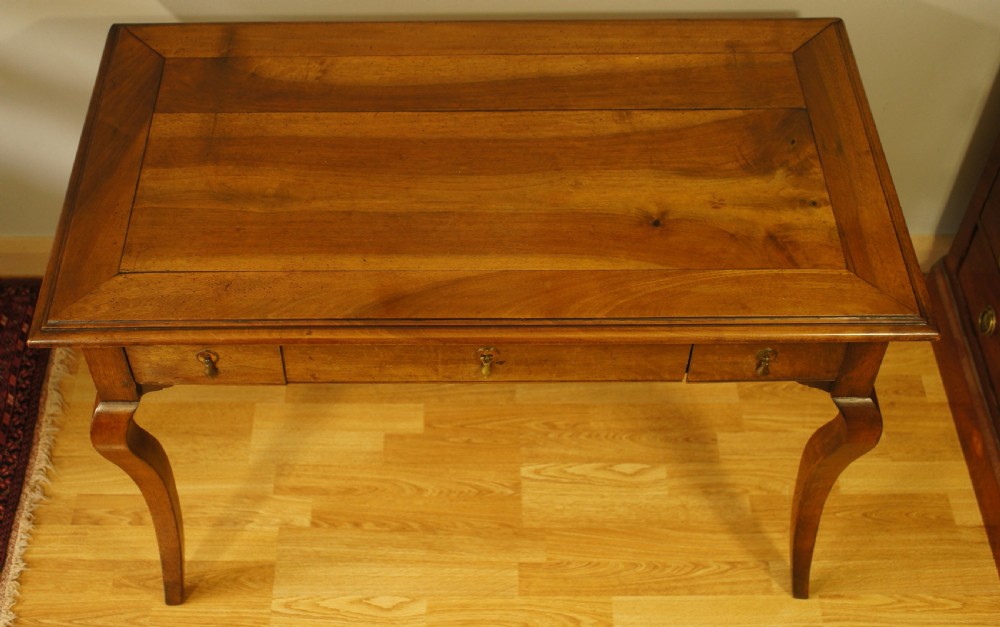 An antique French walnut writing table featuring three frieze drawers, brass handles, and cabriole legs against a carved wooden panelled backdrop. View VI