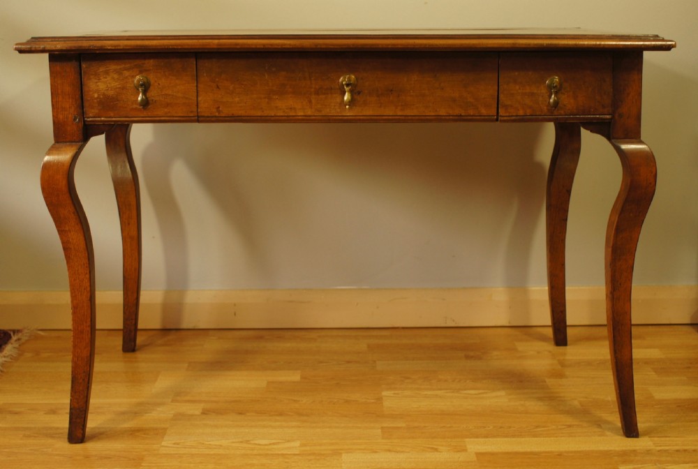 An antique French walnut writing table featuring three frieze drawers, brass handles, and cabriole legs against a carved wooden panelled backdrop. View VIII