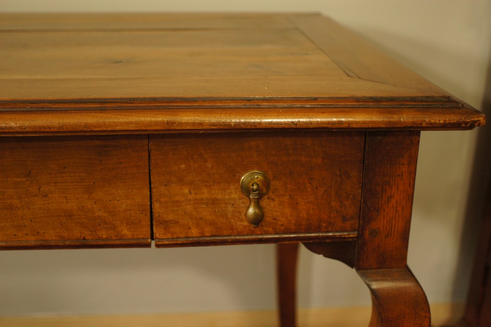 An antique French walnut writing table featuring three frieze drawers, brass handles, and cabriole legs against a carved wooden panelled backdrop. View X