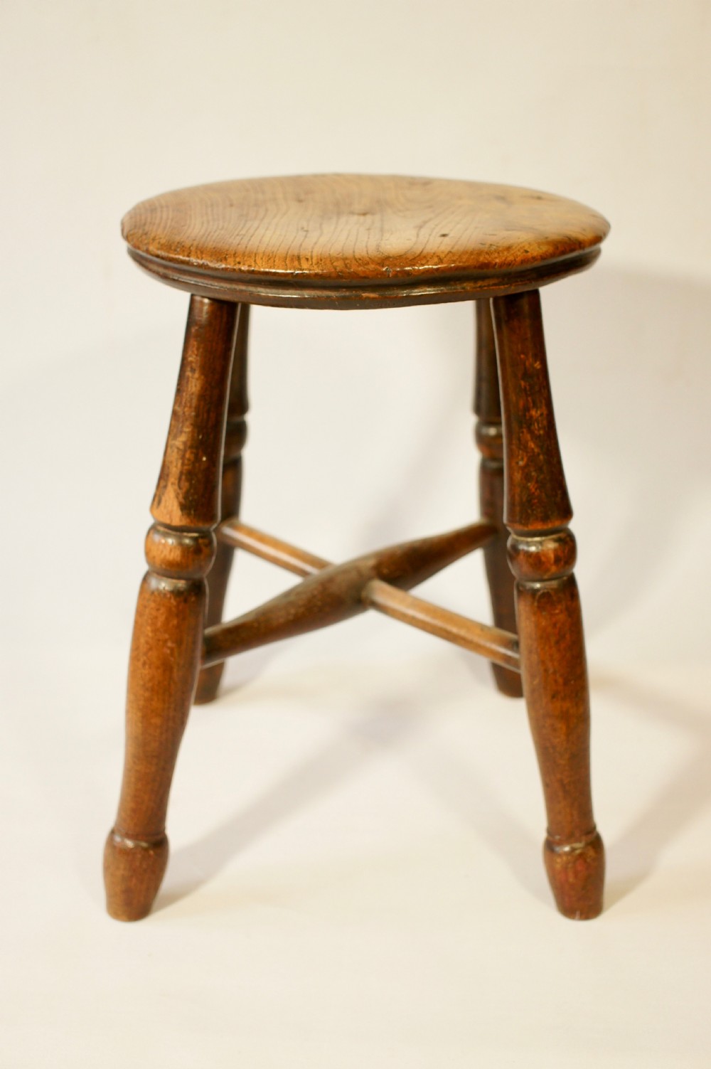 An antique wooden stool featuring a circular solid elm seat resting upon four splayed turned legs joined by an X-frame stretcher.