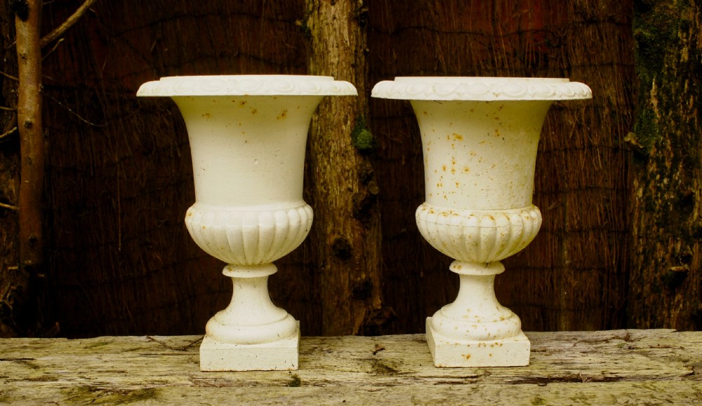 A pair of off-white painted cast iron urns featuring fluted lower bodies, flared upper rims, turned stems, and square plinth bases with visible surface rust.