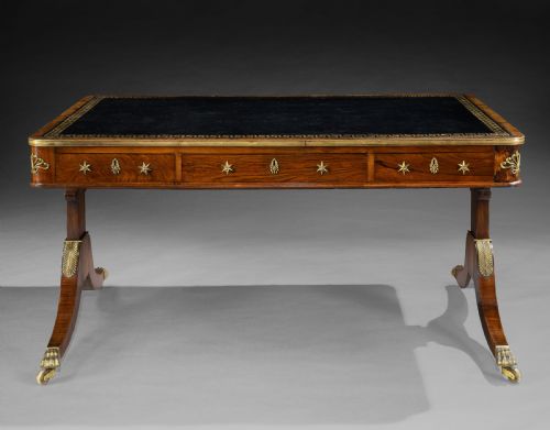 fig 1. A Regency style rosewood writing table featuring a dark leather inset top with gilt Greek key tooling, brass star mounts along the frieze, and trestle legs on brass paw castors.