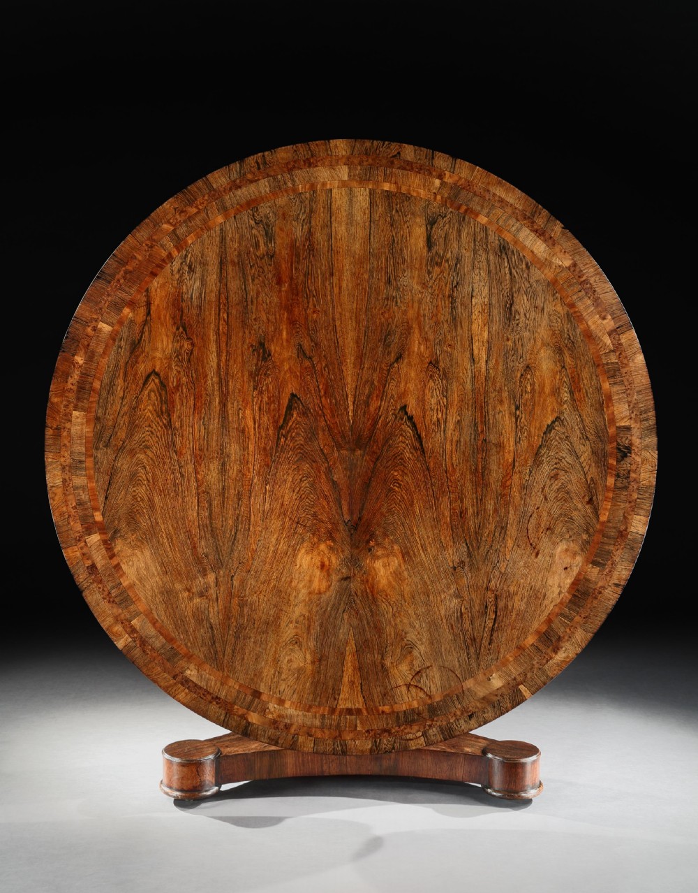 A circular antique rosewood centre table with book-matched grain and crossbanded border shown tilted upright on its base.