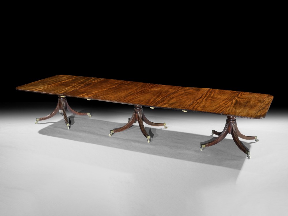 A long George III mahogany dining table featuring three reeded pedestal bases with splayed legs and brass castors.