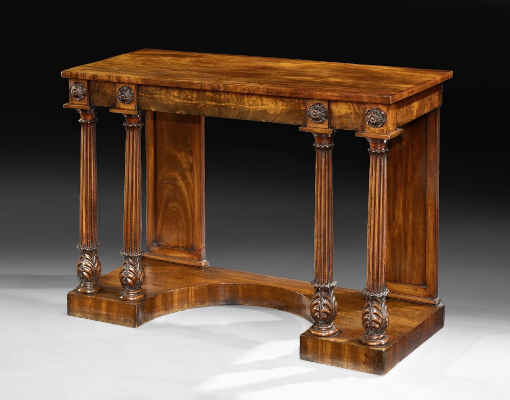 An antique George IV flame mahogany console table featuring twin fluted columns with carved acanthus leaf capitals, floral rosettes on the frieze, and a shaped plinth base.