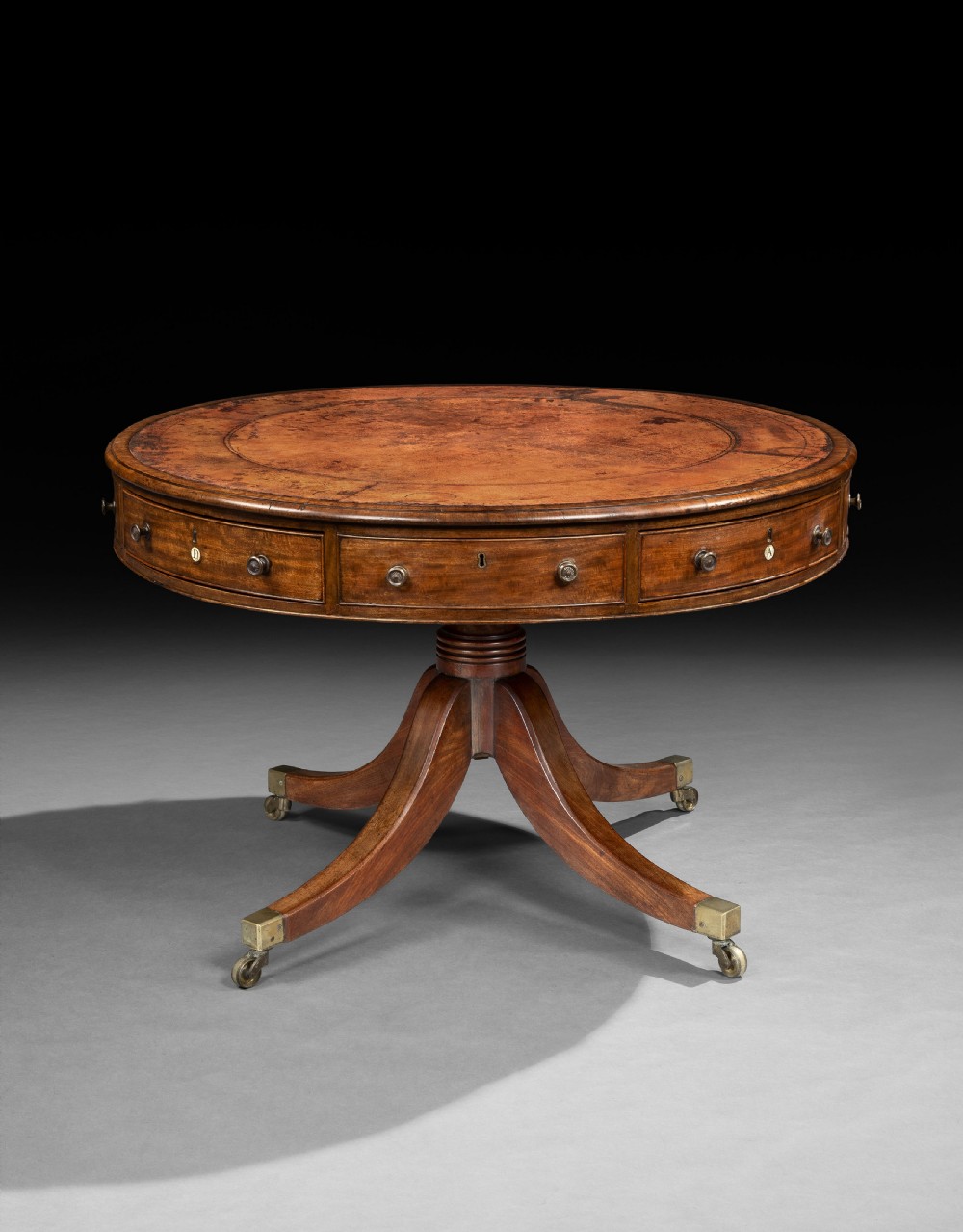 A George III mahogany drum table featuring a circular leather-lined top over frieze drawers, supported on a turned central pedestal with four splayed legs terminating in brass castors.
