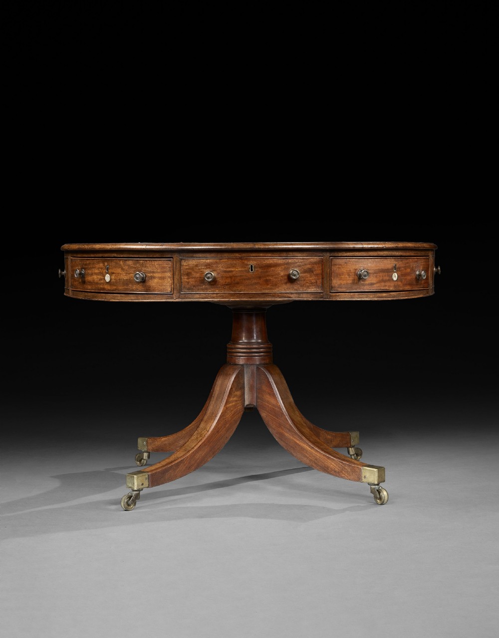 A George III mahogany drum table featuring a circular leather-lined top over frieze drawers, supported on a turned central pedestal with four splayed legs terminating in brass castors. View II
