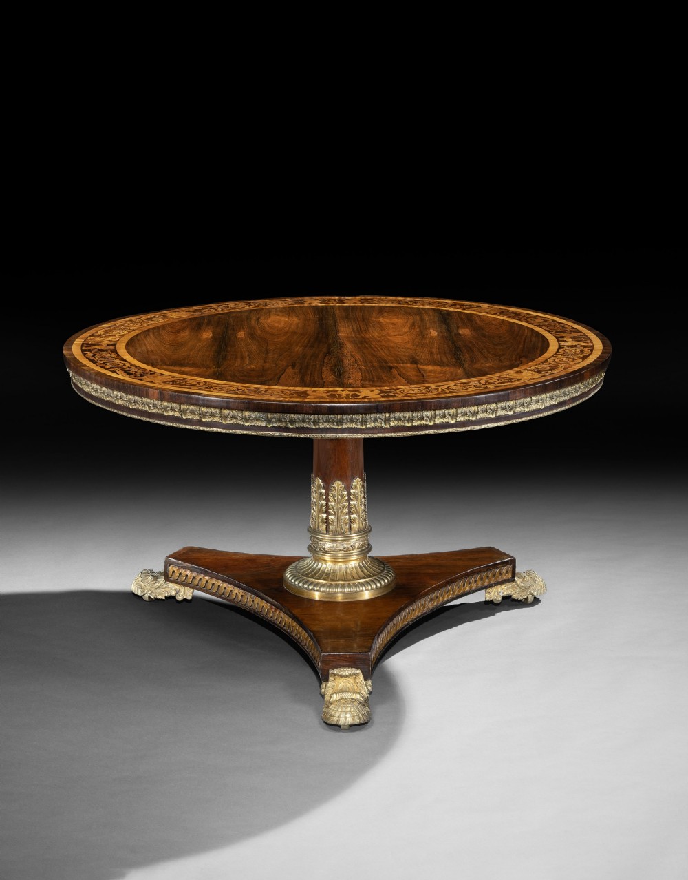 A circular George IV rosewood centre table featuring marquetry inlay along the edge, a brass-mounted pedestal, and a tripartite base on brass paw feet.