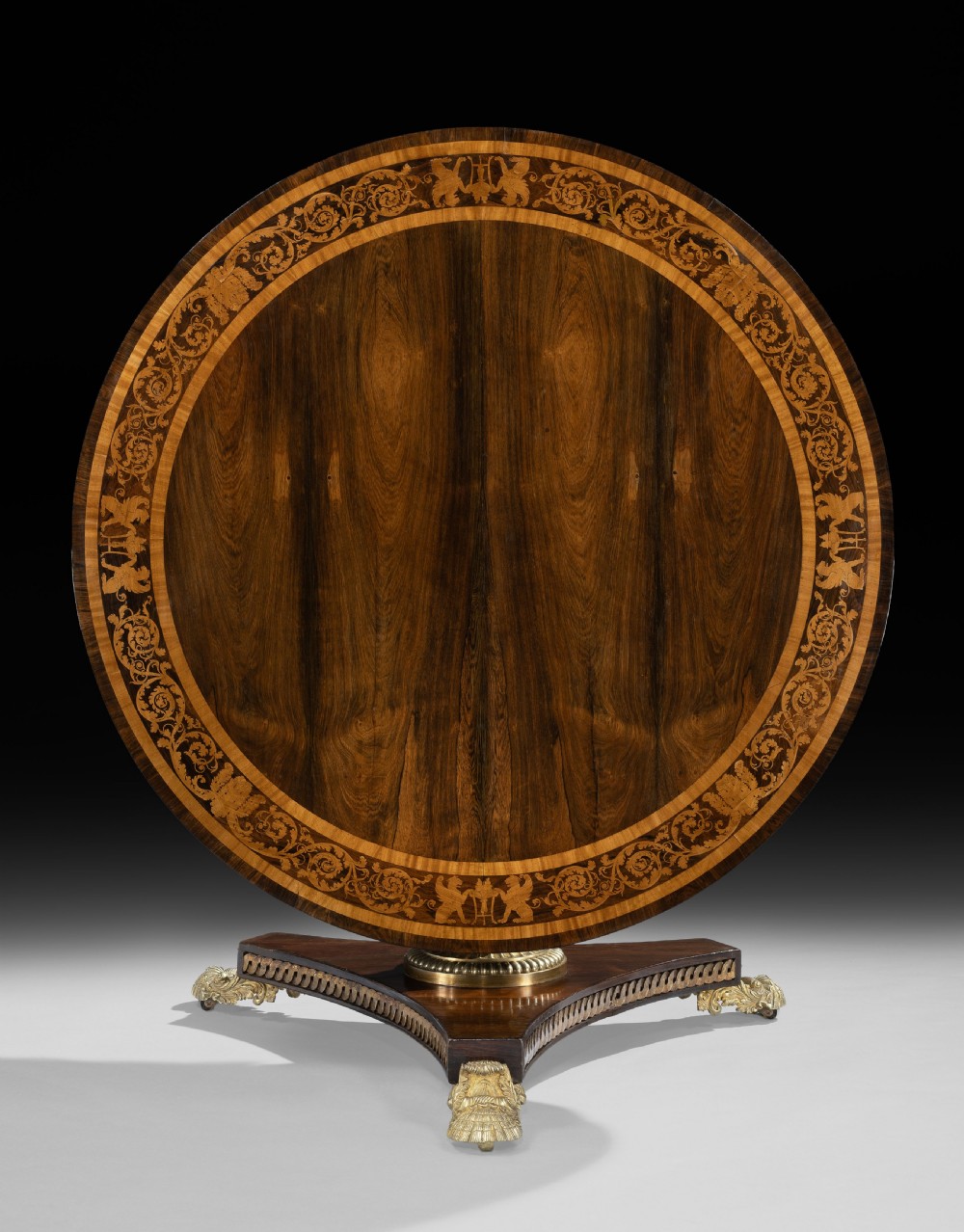 A circular George IV rosewood centre table featuring marquetry inlay along the edge, a brass-mounted pedestal, and a tripartite base on brass paw feet. View II