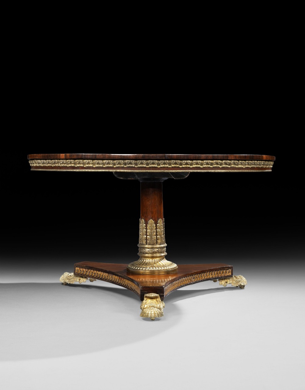 A circular George IV rosewood centre table featuring marquetry inlay along the edge, a brass-mounted pedestal, and a tripartite base on brass paw feet. View III