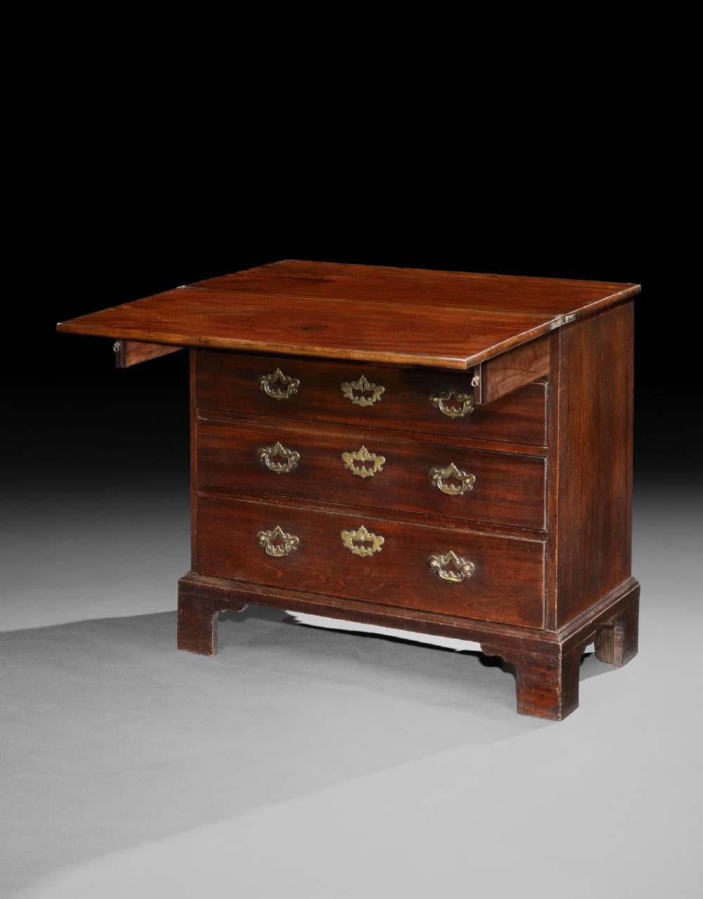 An 18th-century George II mahogany bachelors chest featuring a hinged folding top leaf, three long drawers with shaped brass handles, and bracket feet.