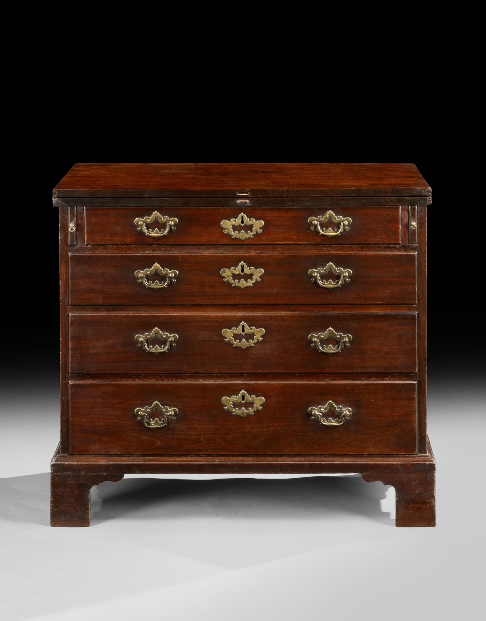 An 18th-century George II mahogany bachelors chest featuring a hinged folding top leaf, three long drawers with shaped brass handles, and bracket feet. View II