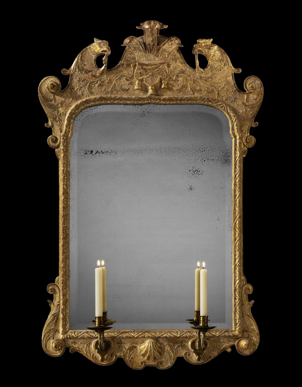 A George I carved giltwood wall mirror featuring an ornate crest with eagle motifs, a lower carved shell apron, and twin brass candle sconces.