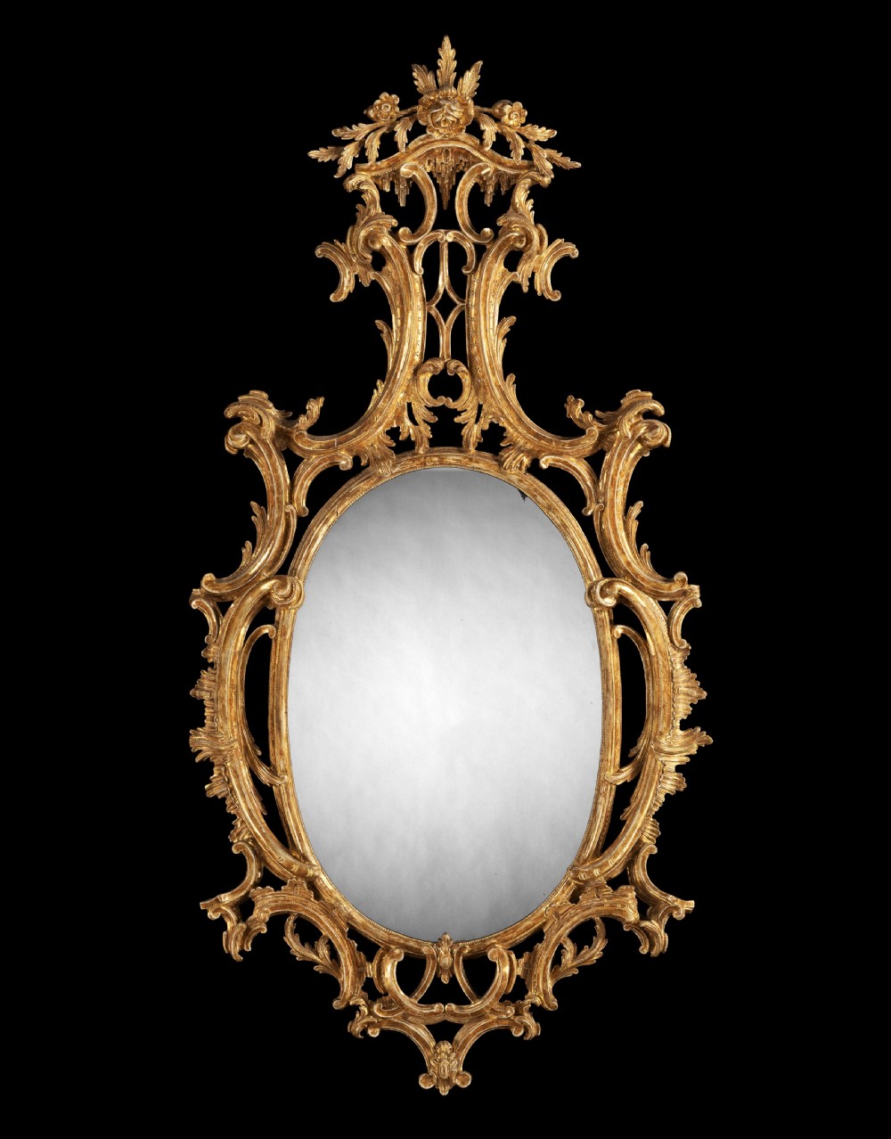 An oval George III giltwood wall mirror featuring an openwork frame with C-scrolls, acanthus leaf carving, and a tall floral crest.