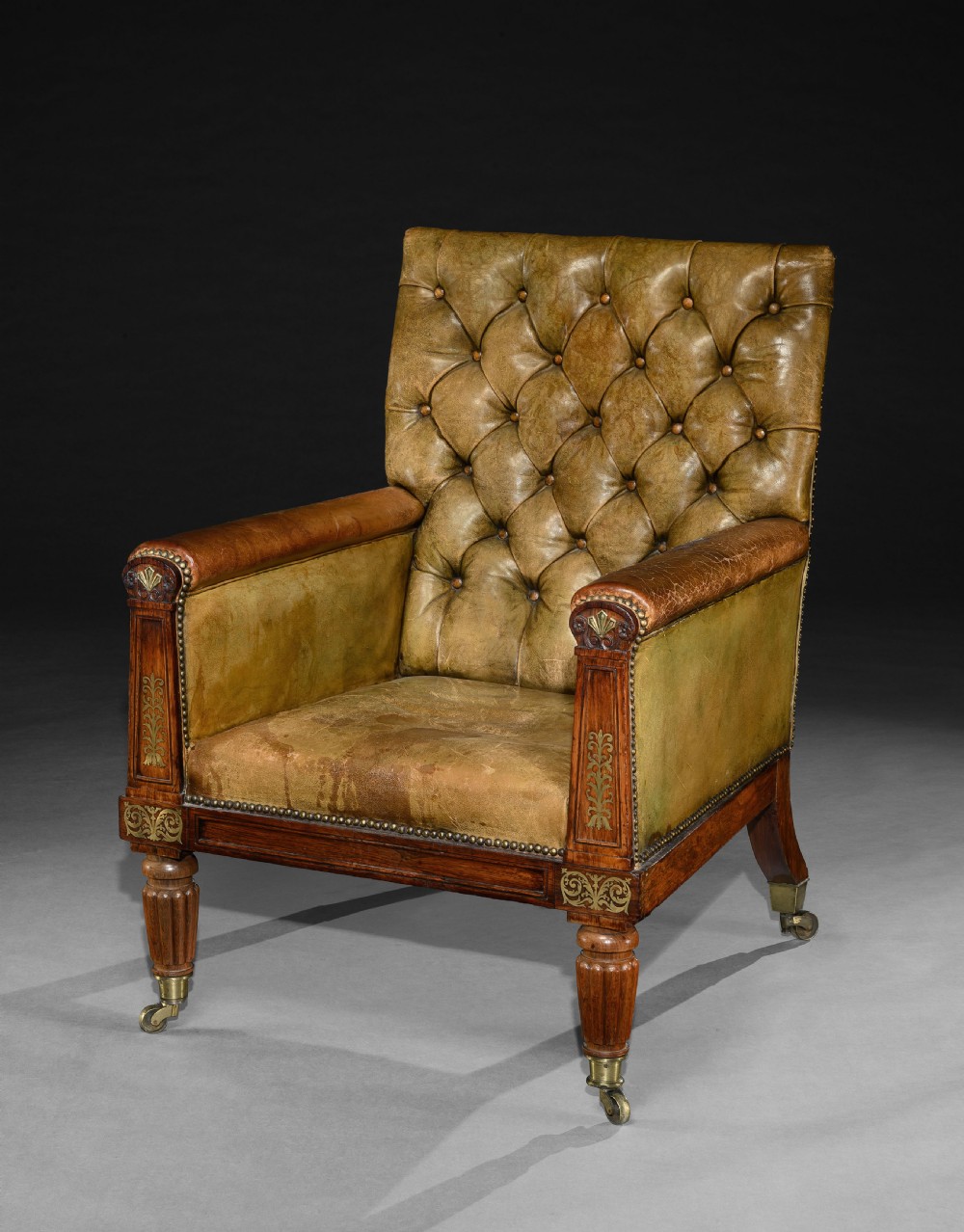 A George IV rosewood library armchair featuring button-tufted green leather upholstery, carved anthemion motifs, brass inlay, and reeded legs with castors.