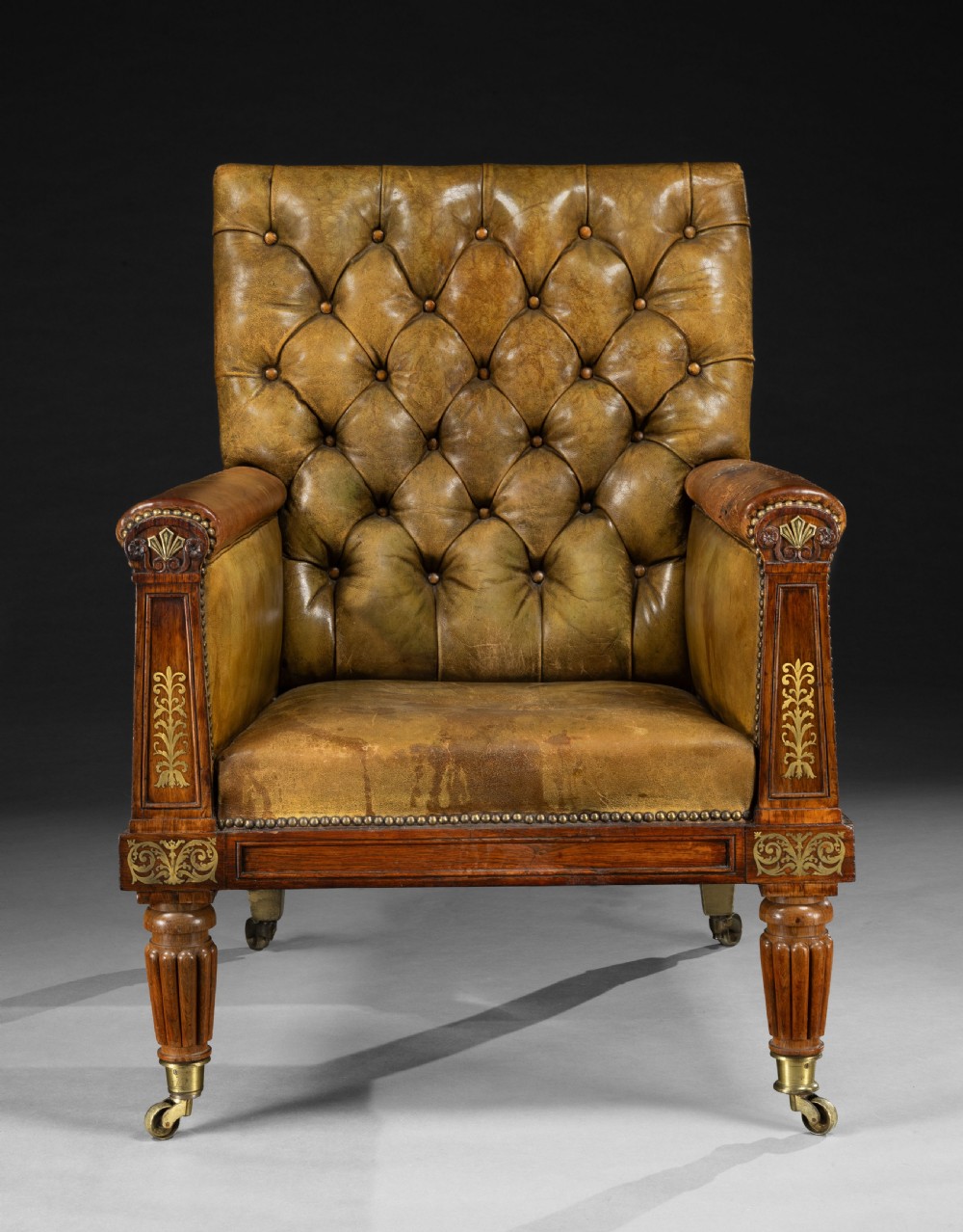 A George IV rosewood library armchair featuring button-tufted green leather upholstery, carved anthemion motifs, brass inlay, and reeded legs with castors. View II