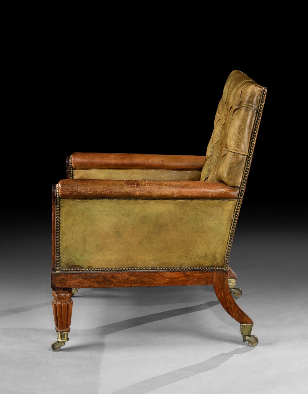 A George IV rosewood library armchair featuring button-tufted green leather upholstery, carved anthemion motifs, brass inlay, and reeded legs with castors. View III
