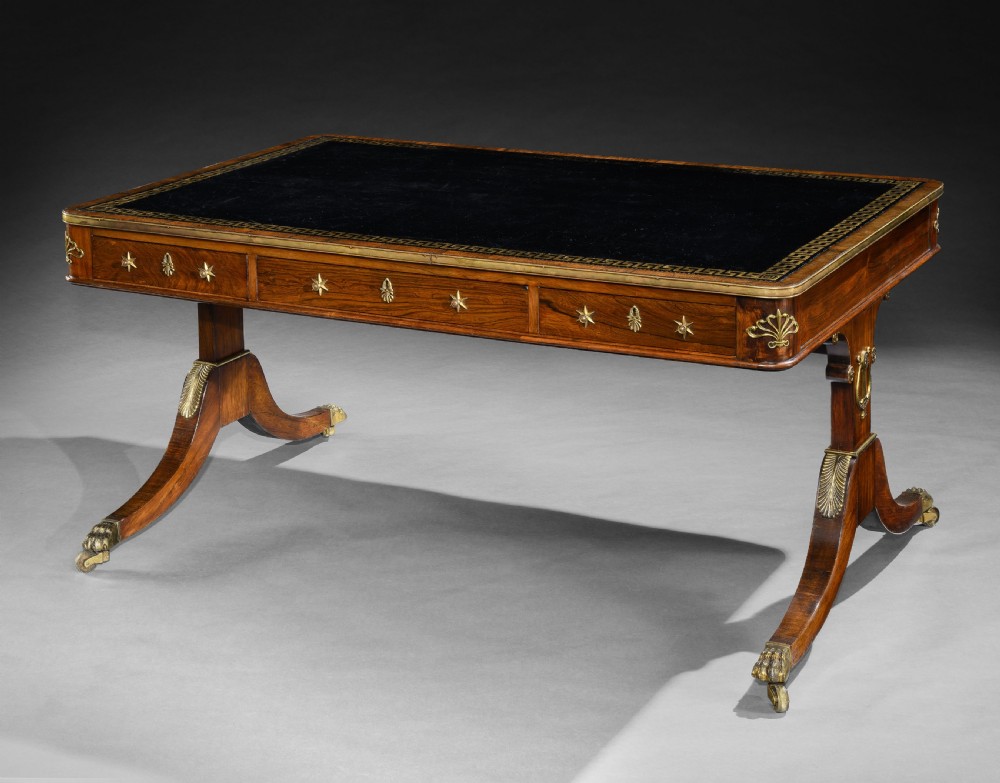 A Regency style rosewood writing table featuring a dark leather inset top with gilt Greek key tooling, brass star mounts along the frieze, and trestle legs on brass paw castors.