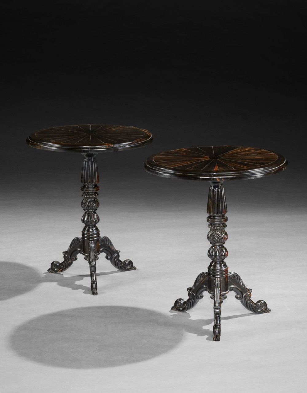 A pair of dark carved ebony tripod tables with radial inlaid circular tops and turned pedestals on scroll feet.