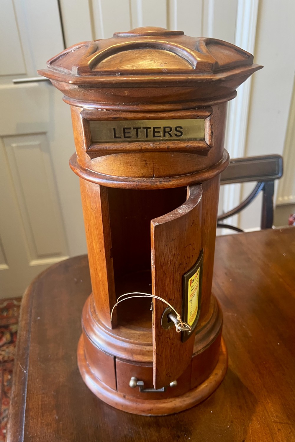 A cylindrical wooden post box featuring a brass letter slot, a small door with a key, and a lower drawer with a metal handle. View VII