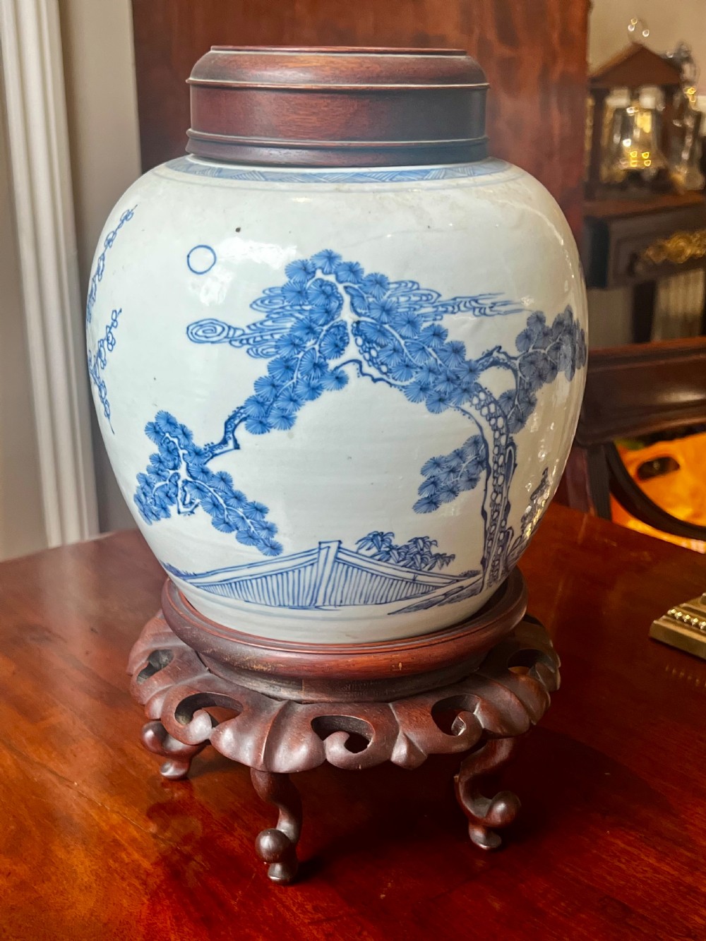 A blue and white porcelain ginger jar featuring hand-painted botanical motifs, complete with a wooden cover and resting on an intricately carved wooden stand.