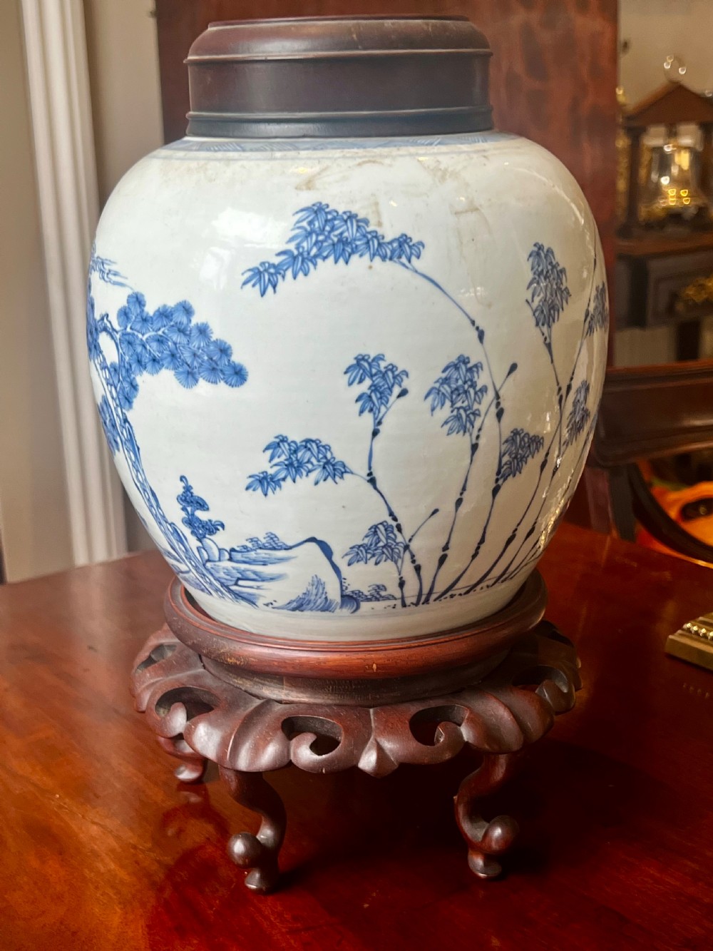 A blue and white porcelain ginger jar featuring hand-painted botanical motifs, complete with a wooden cover and resting on an intricately carved wooden stand. View II