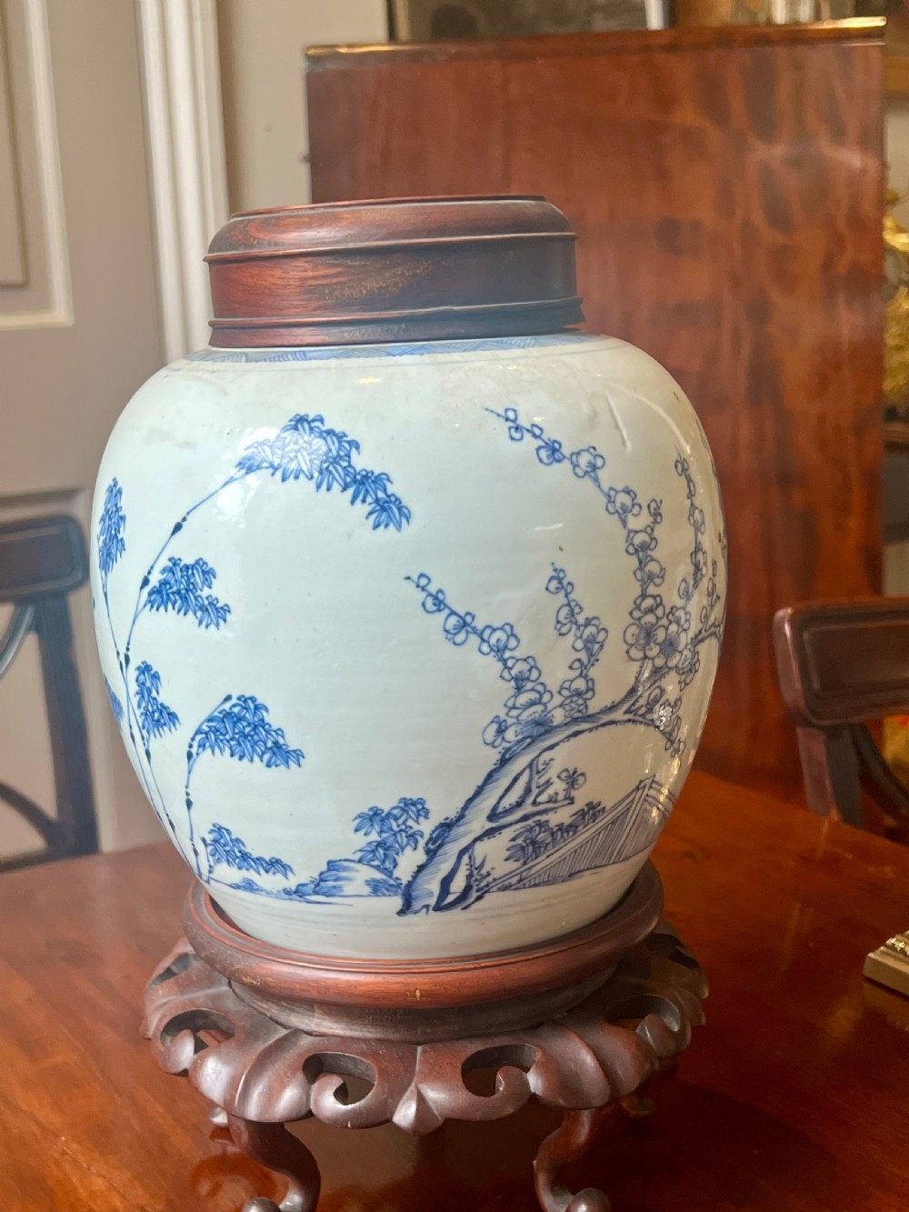 A blue and white porcelain ginger jar featuring hand-painted botanical motifs, complete with a wooden cover and resting on an intricately carved wooden stand. View III
