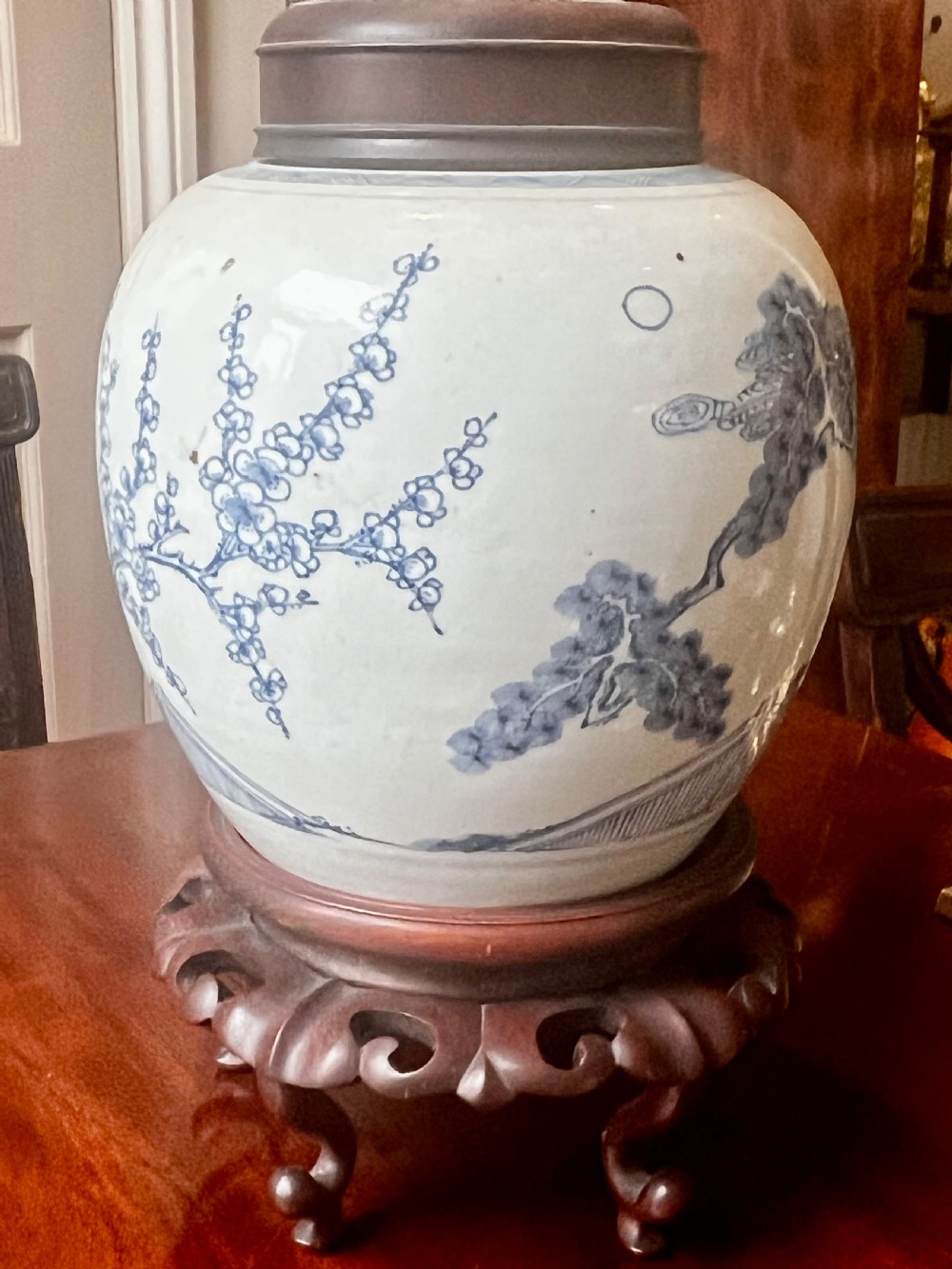 A blue and white porcelain ginger jar featuring hand-painted botanical motifs, complete with a wooden cover and resting on an intricately carved wooden stand. View VI