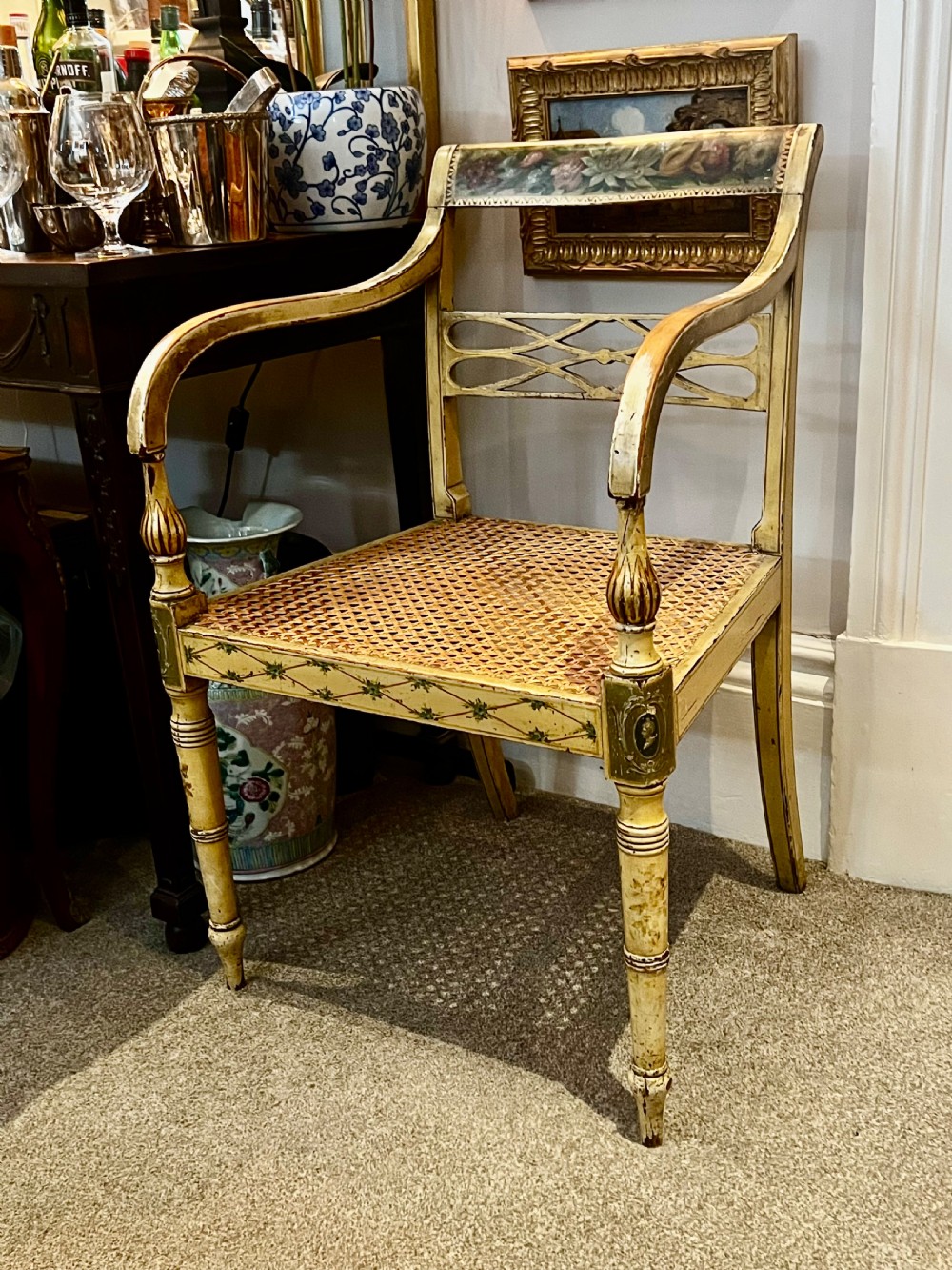 A Regency period cream painted elbow chair featuring hand-painted crest rail decoration, a geometric lattice back, sweeping arms, turned front legs, and a woven cane seat.