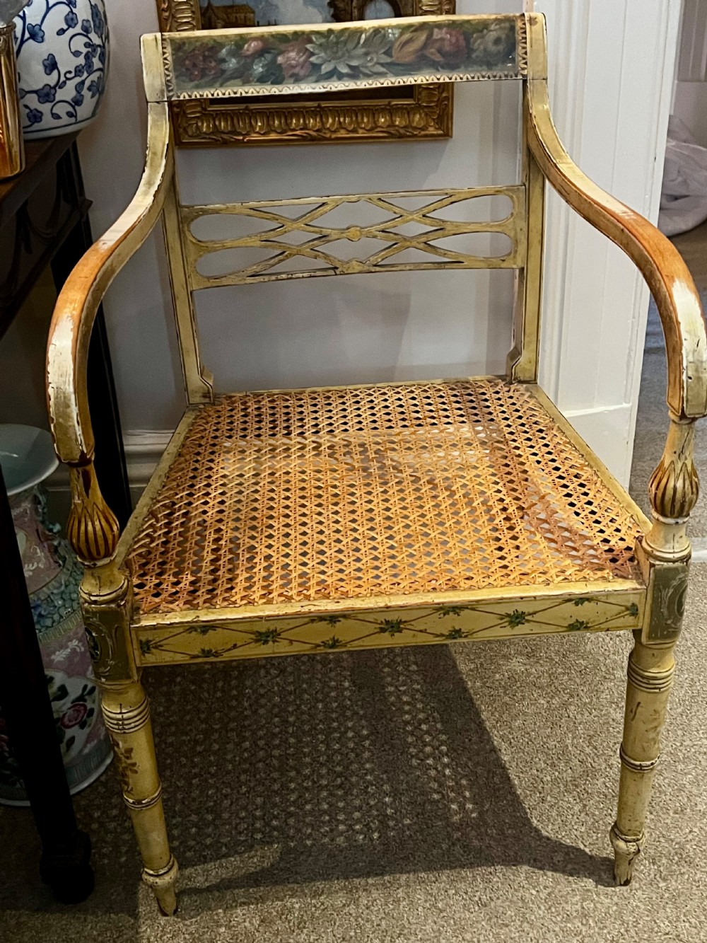 A Regency period cream painted elbow chair featuring hand-painted crest rail decoration, a geometric lattice back, sweeping arms, turned front legs, and a woven cane seat. View IV