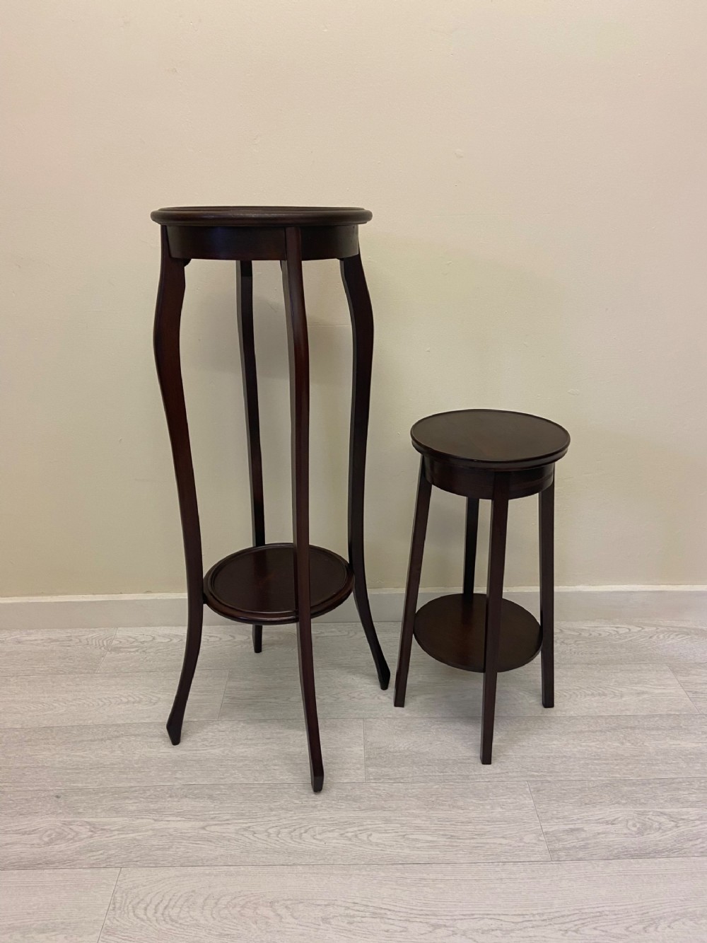 A non-matching pair of dark brown mahogany plant stands featuring circular tops, lower undertiers, and curved legs.