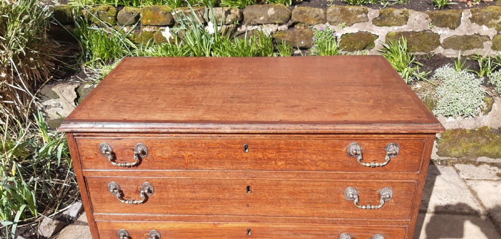 A Georgian oak chest of drawers featuring four full-width drawers with brass handles and keyhole escutcheons, resting on bracket feet. View V
