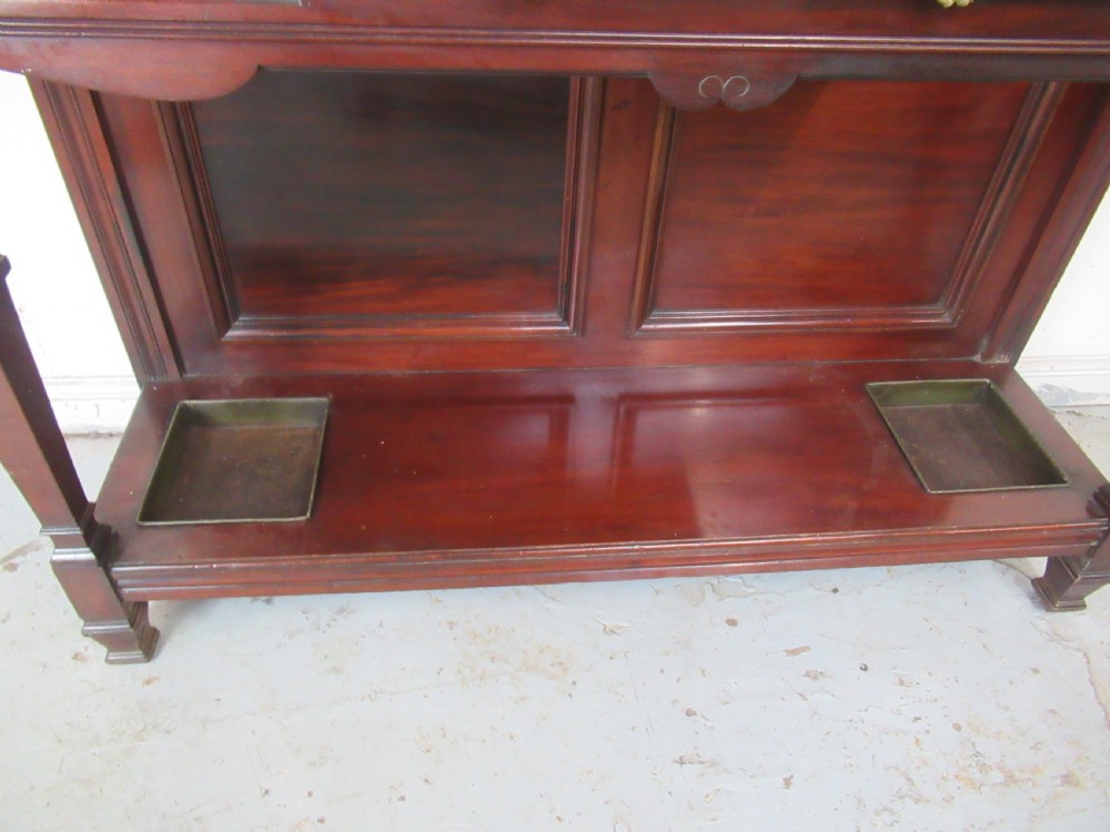 A tall Victorian mahogany hall stand featuring a central rectangular mirror, carved wooden panels, brass coat hooks, a central drawer, and integrated umbrella stands with drip trays. View III