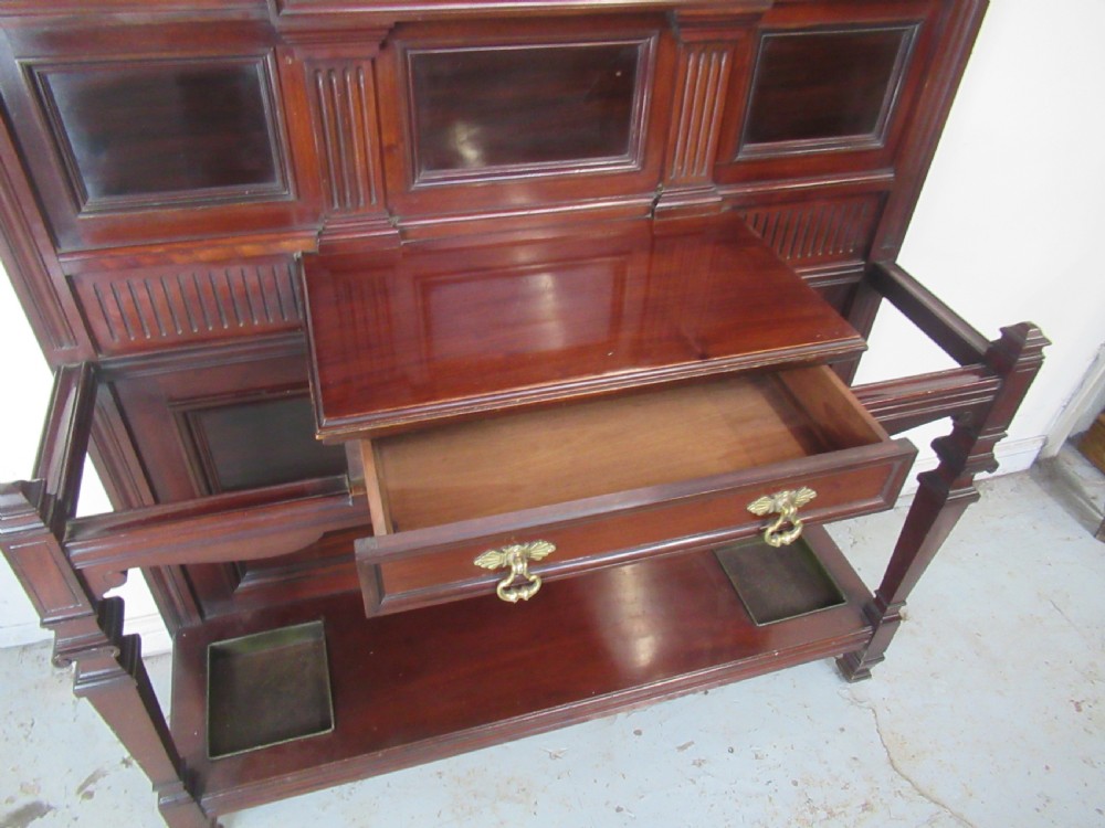 A tall Victorian mahogany hall stand featuring a central rectangular mirror, carved wooden panels, brass coat hooks, a central drawer, and integrated umbrella stands with drip trays. View V