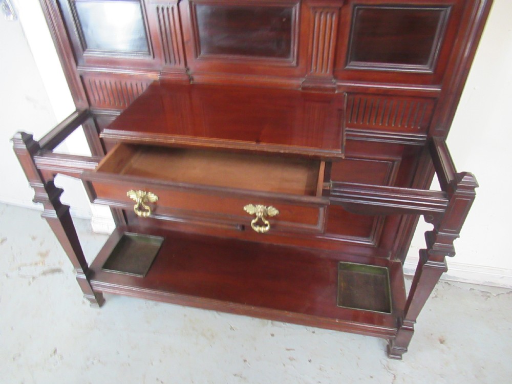 A tall Victorian mahogany hall stand featuring a central rectangular mirror, carved wooden panels, brass coat hooks, a central drawer, and integrated umbrella stands with drip trays. View VI
