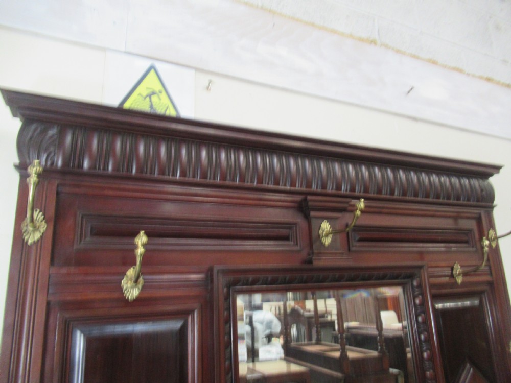 A tall Victorian mahogany hall stand featuring a central rectangular mirror, carved wooden panels, brass coat hooks, a central drawer, and integrated umbrella stands with drip trays. View IX