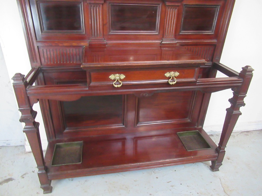 A tall Victorian mahogany hall stand featuring a central rectangular mirror, carved wooden panels, brass coat hooks, a central drawer, and integrated umbrella stands with drip trays. View X