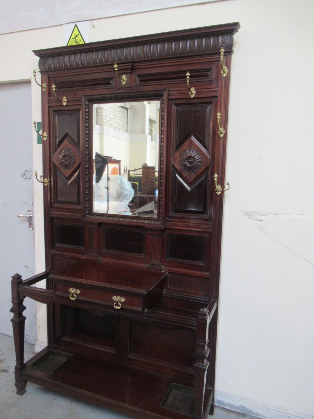 A tall Victorian mahogany hall stand featuring a central rectangular mirror, carved wooden panels, brass coat hooks, a central drawer, and integrated umbrella stands with drip trays.