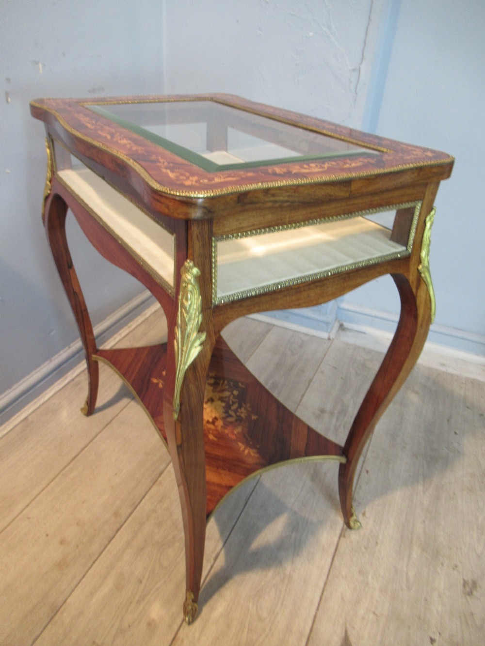 A 19th-century French rosewood bijouterie table featuring a glazed lift top, floral marquetry inlay, gilt bronze mounts, cabriole legs, and a lower shelf.