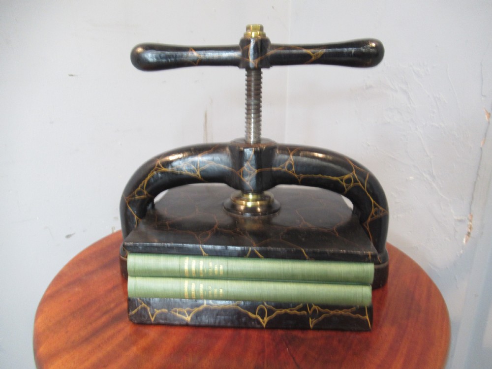 A dark, crackle-finished cast iron and brass book press sits on a round wooden table, clamping two green books.