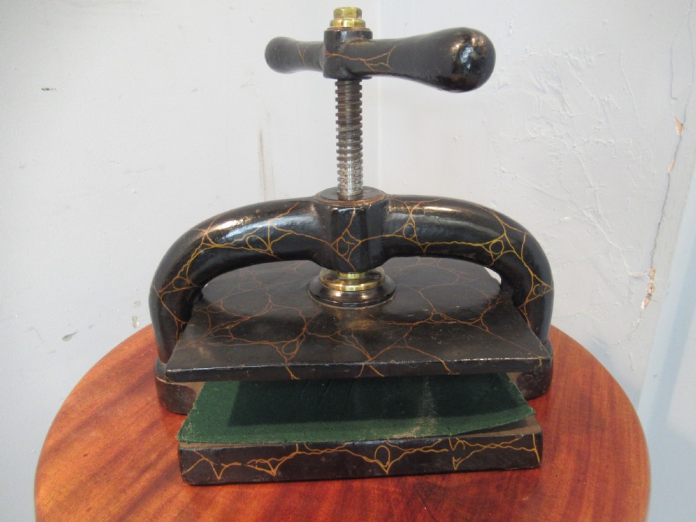 A dark, crackle-finished cast iron and brass book press sits on a round wooden table, clamping two green books. View II