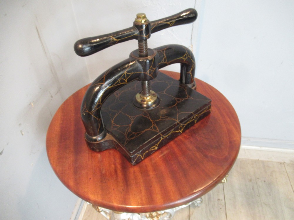A dark, crackle-finished cast iron and brass book press sits on a round wooden table, clamping two green books. View III