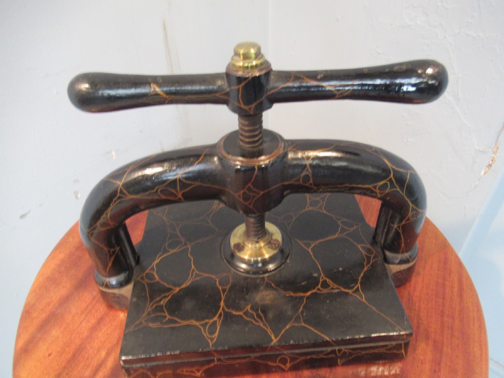 A dark, crackle-finished cast iron and brass book press sits on a round wooden table, clamping two green books. View IV