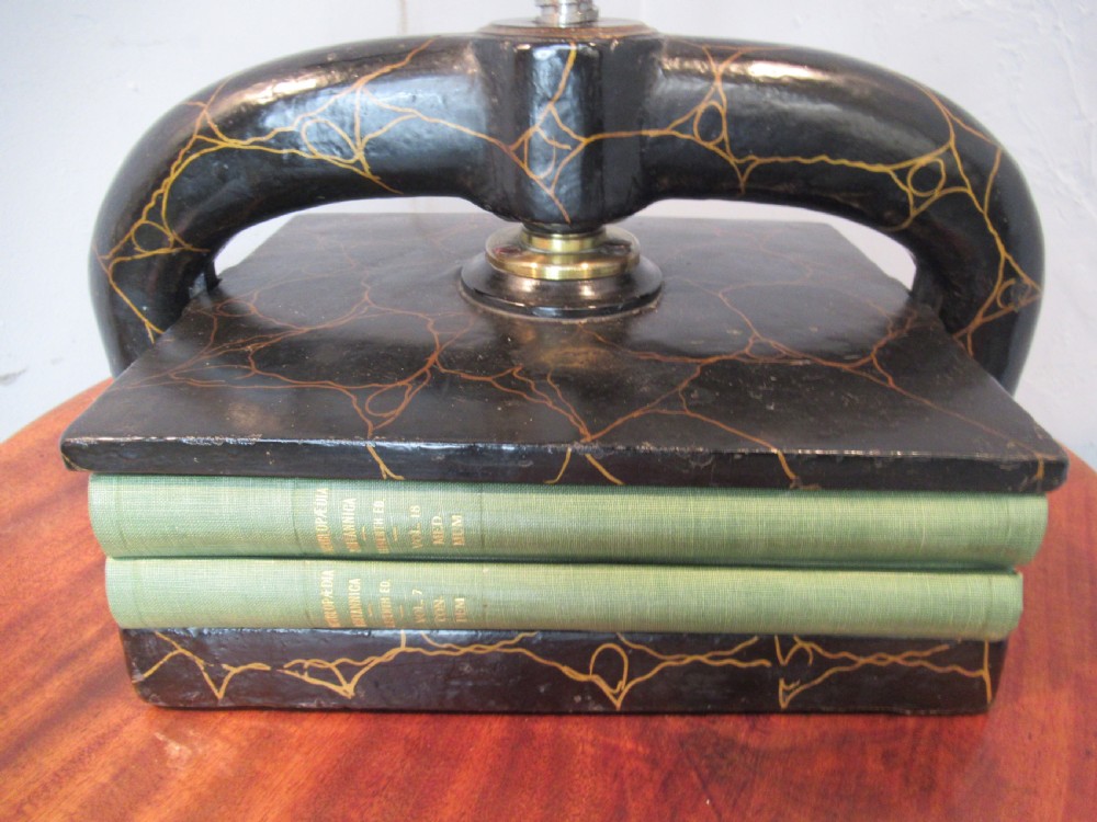 A dark, crackle-finished cast iron and brass book press sits on a round wooden table, clamping two green books. View V