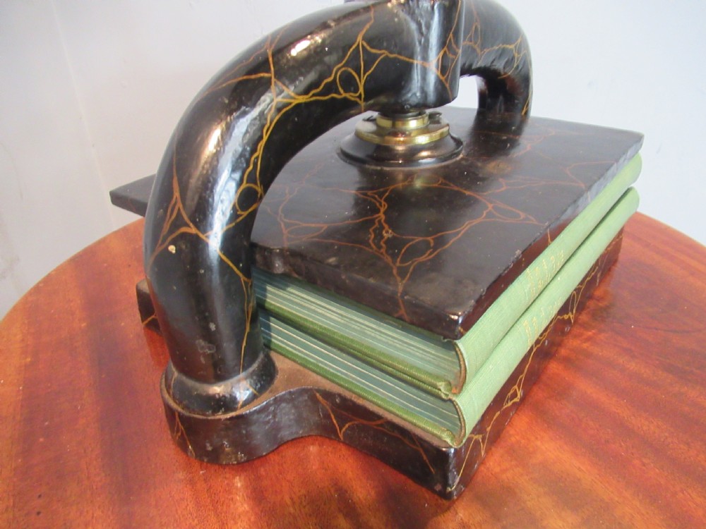 A dark, crackle-finished cast iron and brass book press sits on a round wooden table, clamping two green books. View VI