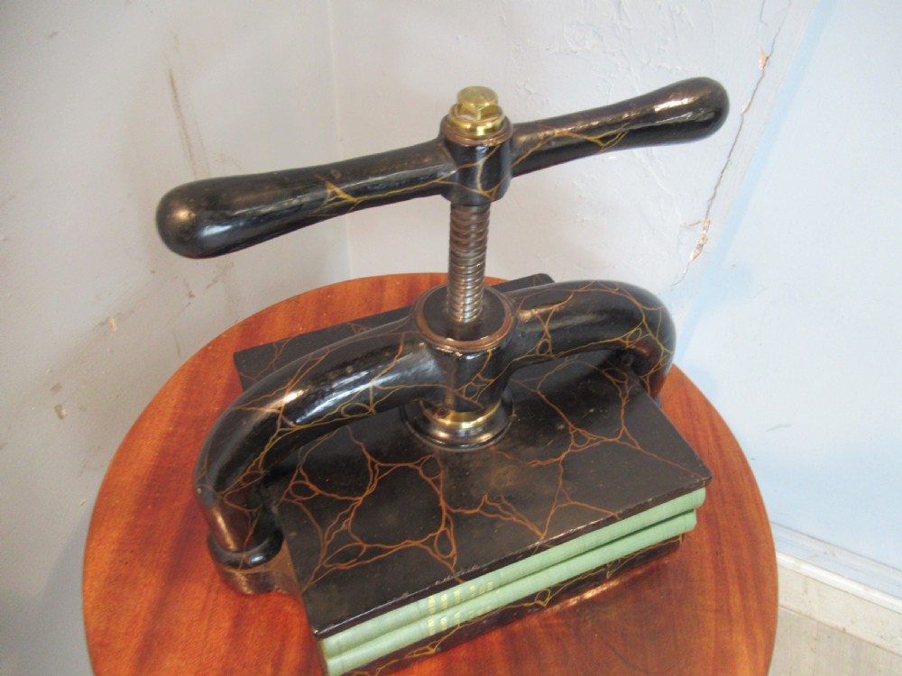 A dark, crackle-finished cast iron and brass book press sits on a round wooden table, clamping two green books. View VII